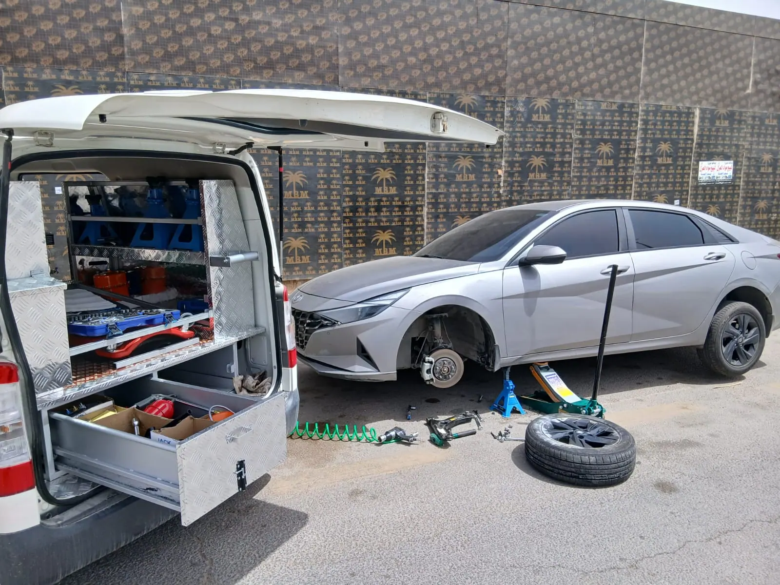 Saaed Service van with its tool drawers open beside a Hyundai Elantra raised on jack stands with a wheel removed, on a Riyadh street