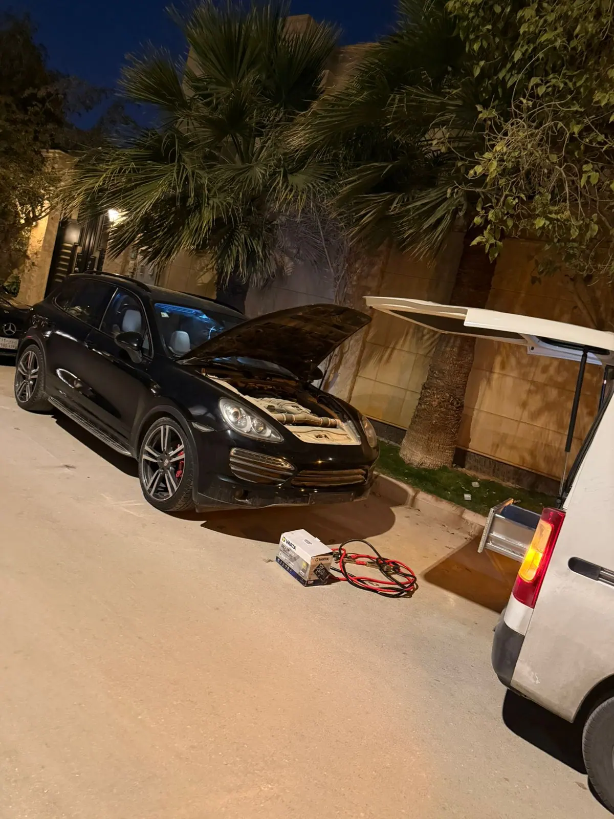 Night battery replacement on a black Porsche Cayenne in Riyadh, with a new battery and jump leads beside the Saaed van